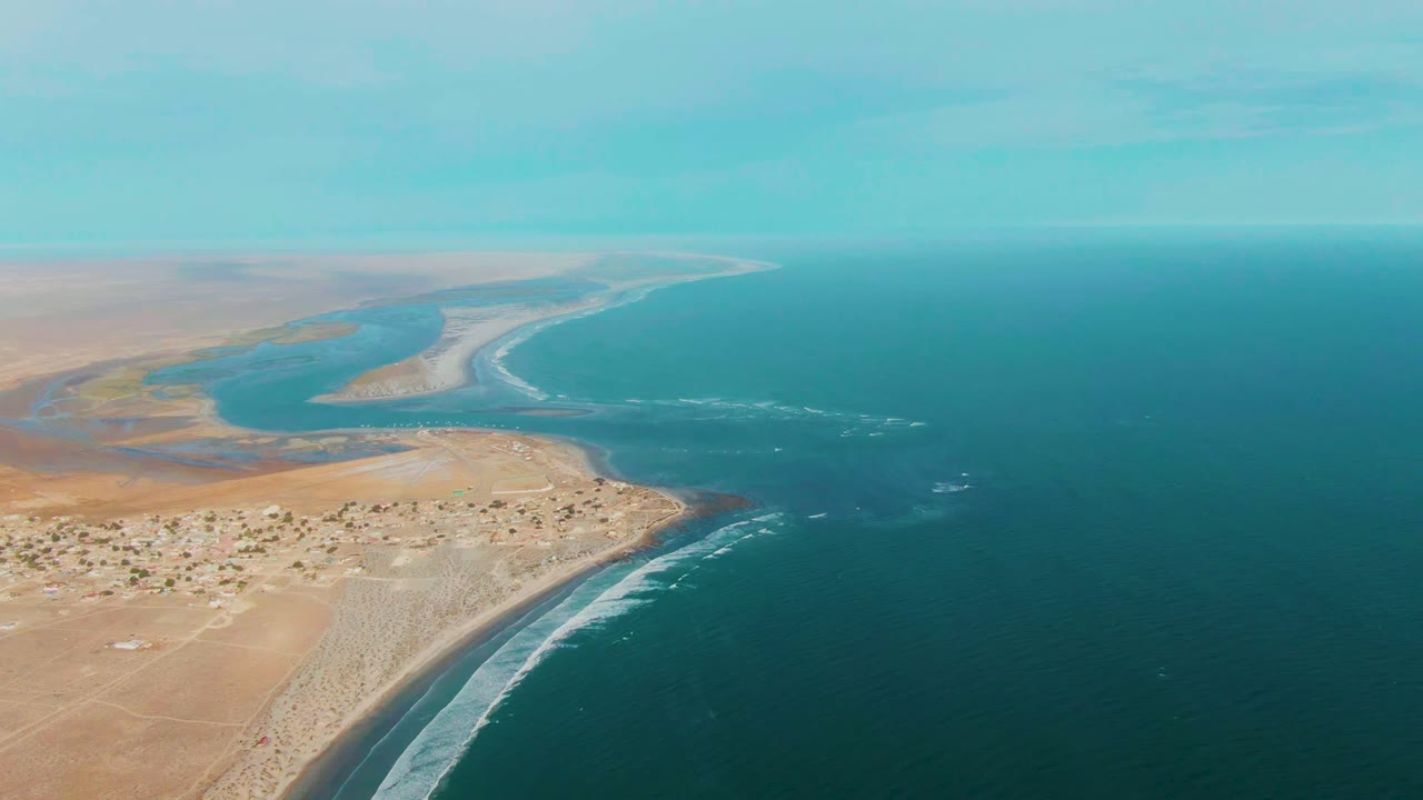 Aerial frame over La Bocana, Baja California Sur, showing a sandy desert shoreline, a river channel entering the ocean, distant low buildings, white surf, and no visible people.