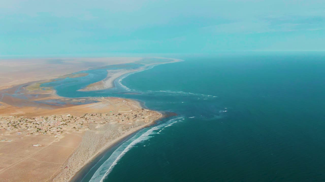 Wide aerial frame at La Bocana with deep blue water on the right, pale desert coast on the left, a river inlet in the distance, and no visible people.