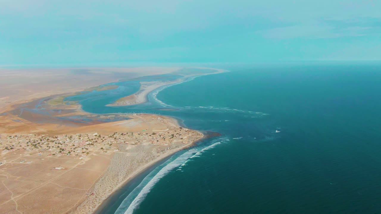 Aerial frame over La Bocana, Baja California Sur showing tan desert land, a small coastal settlement, a curving shoreline, blue ocean, and no visible people.