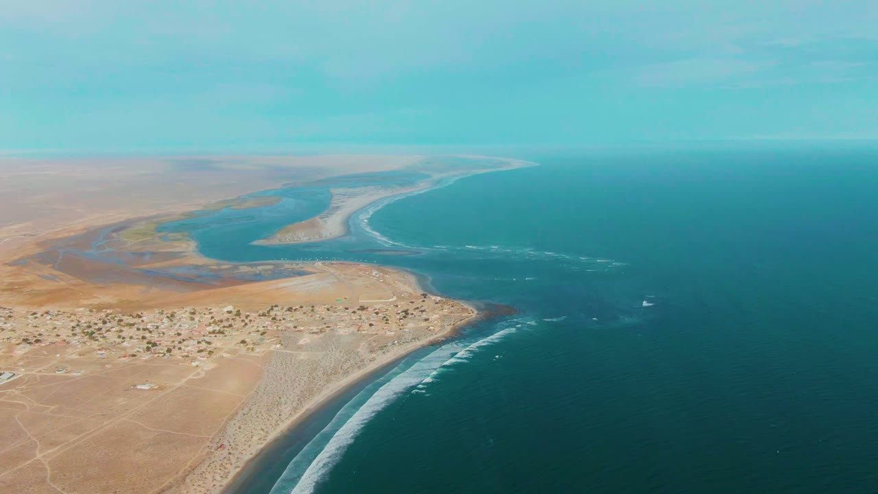 Wide drone view of sandy coast, ocean, settlement, and pale road traces.