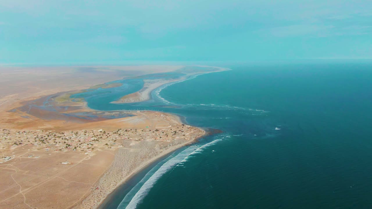 Drone-like aerial frame from La Bocana showing turquoise water, pale surf, desert flats, small buildings near the shore, and no visible people.