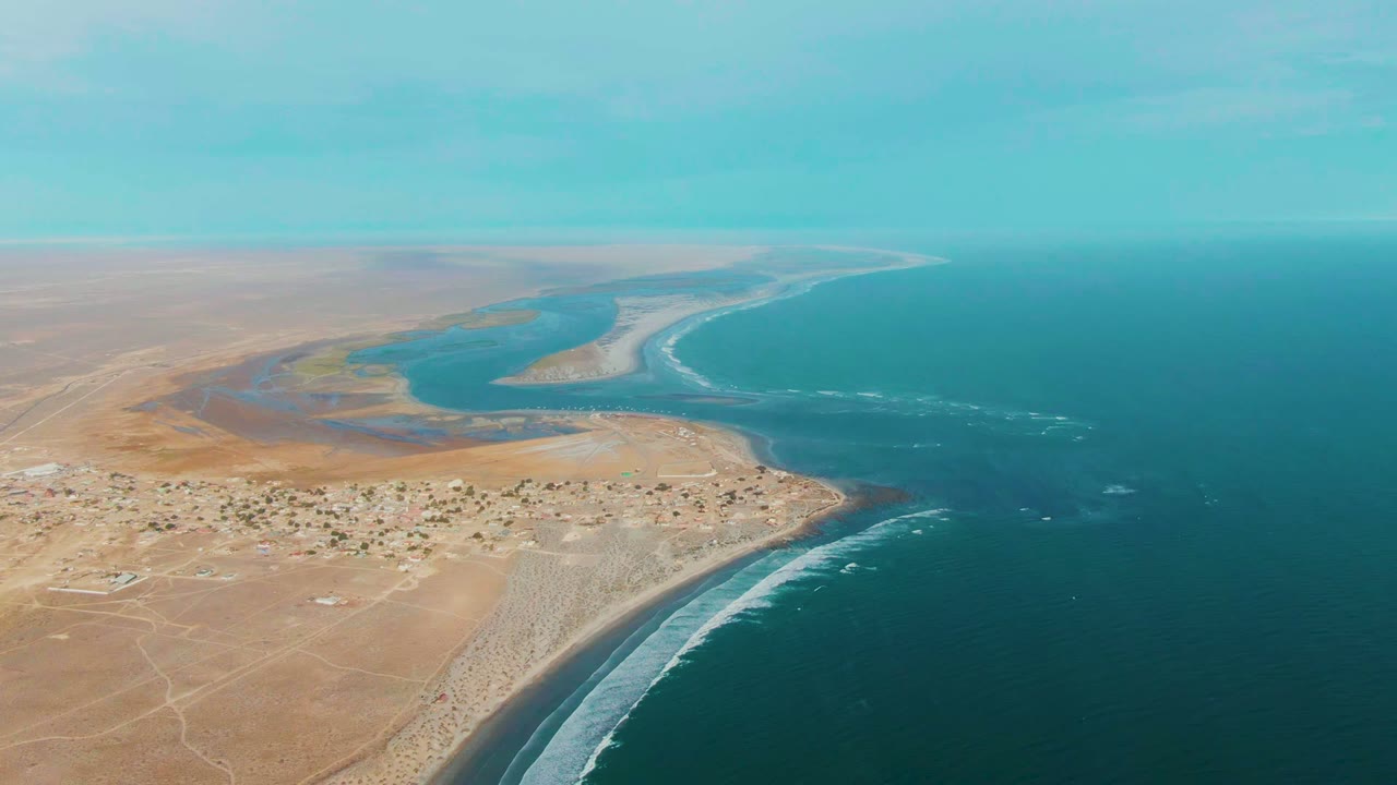 Opening aerial frame over La Bocana, Baja California Sur, Mexico, showing tan desert land, scattered settlement buildings, an estuary, blue ocean, white surf, and no visible people.