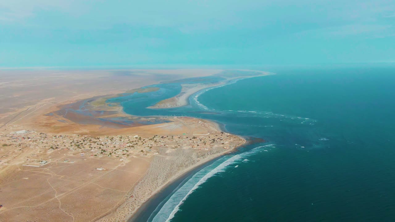 Mid-clip aerial frame over La Bocana with pale desert, clustered small buildings, lagoon-like estuary water, a long surf line, blue ocean, and no visible people.