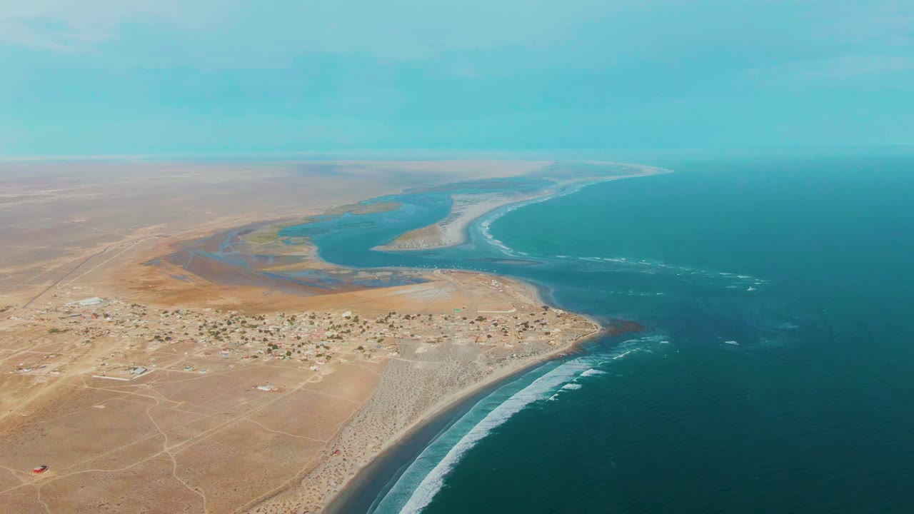 Aerial frame over La Bocana, Baja California Sur showing turquoise lagoon water, sandy desert shoreline, dark ocean, pale surf bands, hazy sky, and no visible people.
