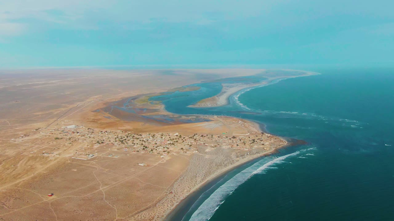Aerial frame over La Bocana, Baja California Sur showing tan desert land, a small settlement, a river estuary, blue ocean, white surf along the shore, and no visible people.
