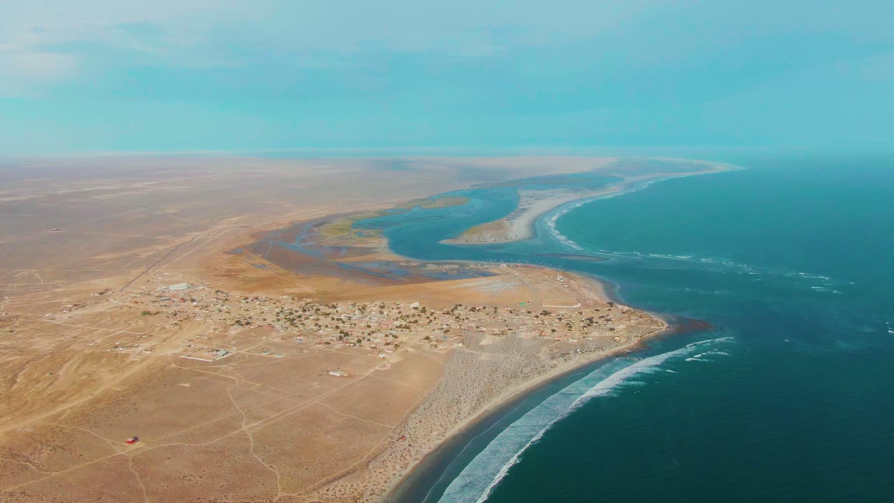 Mid-clip aerial frame of La Bocana, Baja California Sur with the settlement below, a curved river mouth beyond it, blue ocean to the right, white wave bands, and no visible people.