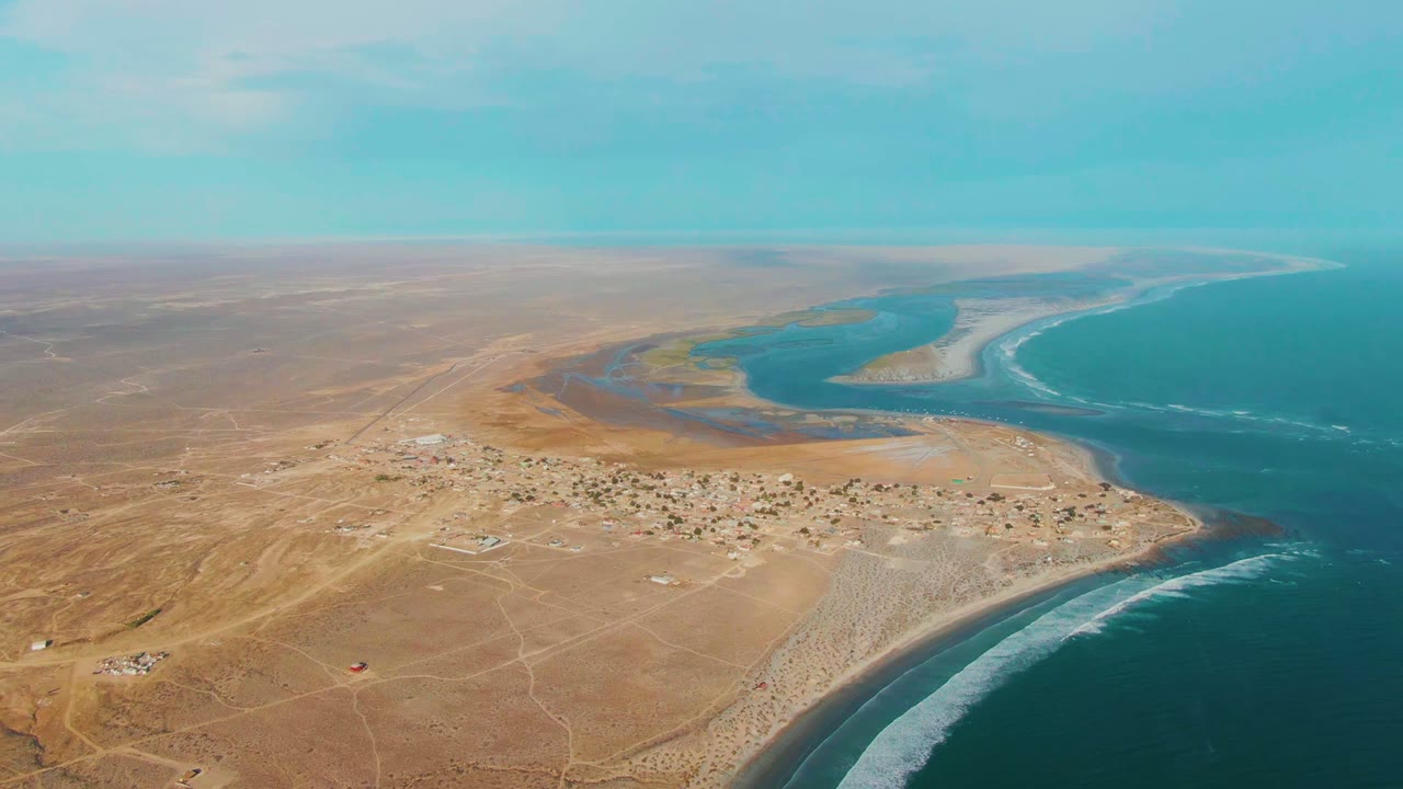 Aerial frame over La Bocana, Baja California Sur, showing tan desert terrain, a small village, curved beach, blue ocean, lagoon water, and no visible people.