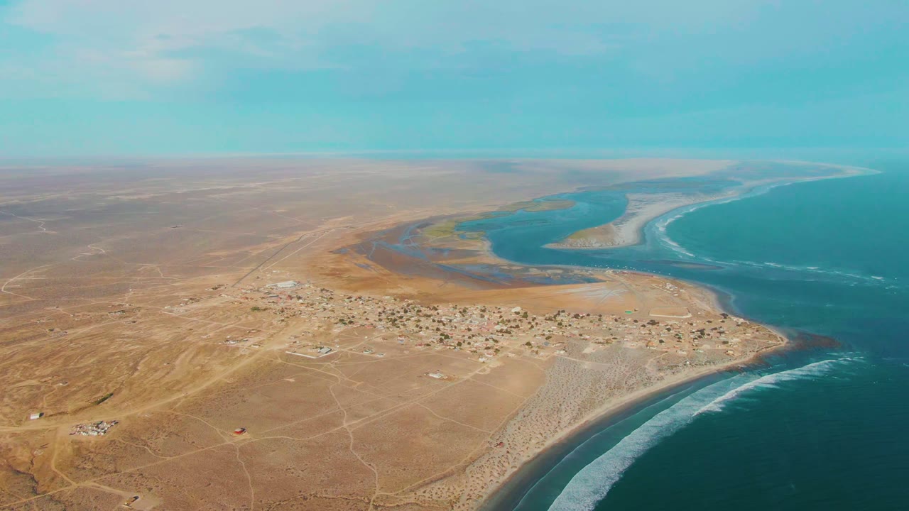 Wide drone view of sandy terrain, settlement, surf line, ocean, and lagoon.