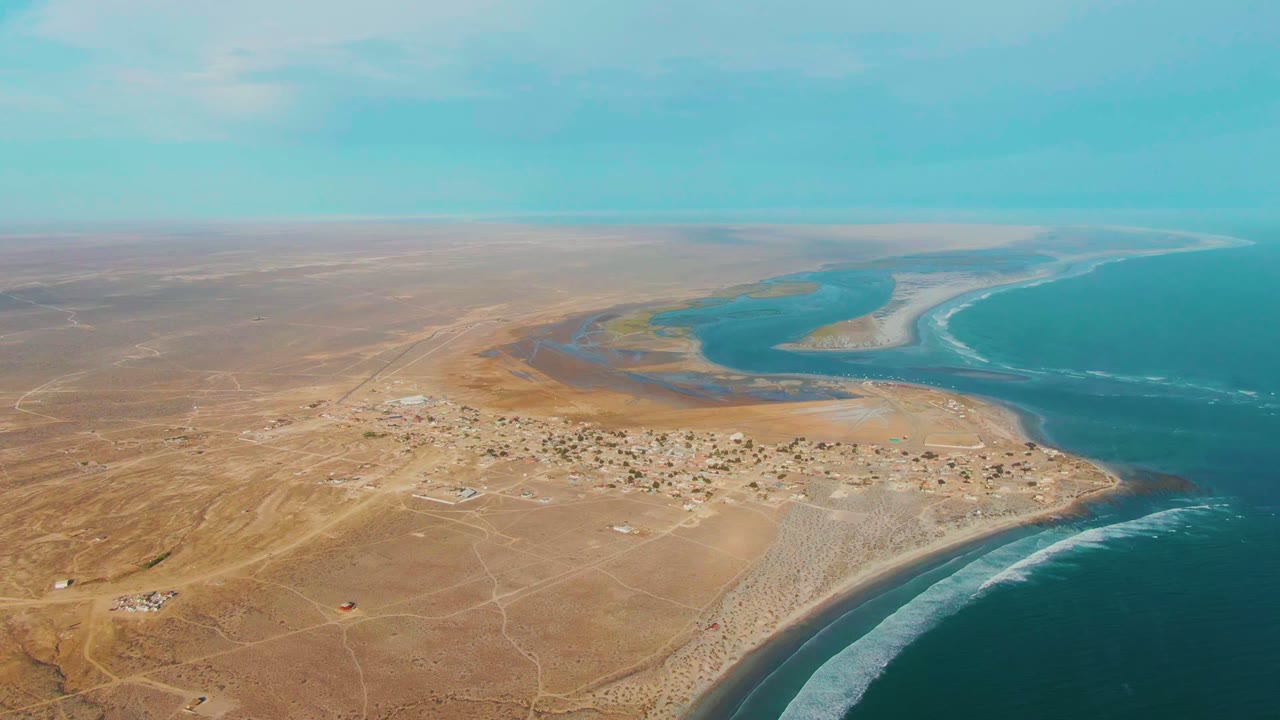 High drone frame over La Bocana showing arid land, a compact settlement, pale roads, dark blue ocean water, white surf, lagoon channels, and no visible people.