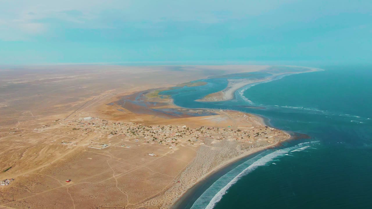 High aerial view over La Bocana, Baja California Sur, with tan desert foreground, a small settlement near the shore, blue ocean to the right, and no visible people.