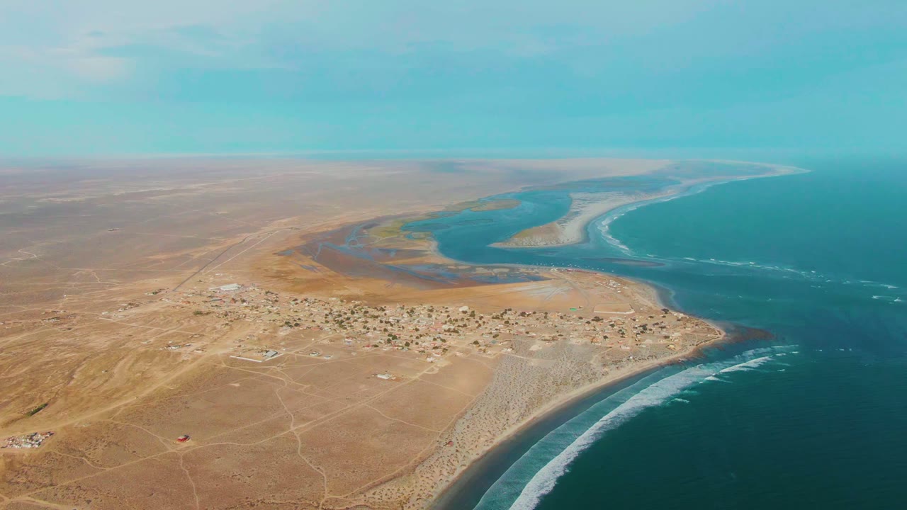 High drone view of La Bocana with a compact settlement near the lower center, a curved river mouth or lagoon behind it, ocean surf on the right, and no visible people.