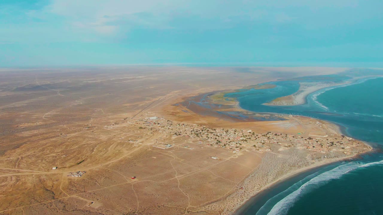 Aerial view over La Bocana, Baja California Sur, with a small desert village, dirt roads, a curving blue inlet, ocean surf, and no visible people.