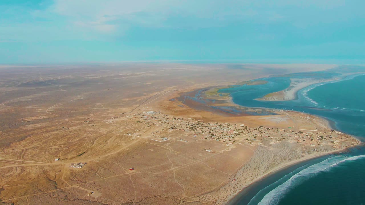 Aerial frame over La Bocana with the village centered below, dry plains stretching inland, a turquoise inlet beyond, ocean surf along the right edge, and no visible people.