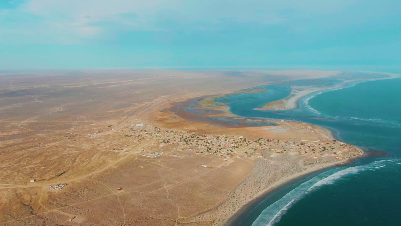 Wide aerial frame over La Bocana, Baja California Sur, with dry desert roads in front, a small settlement near the lagoon, ocean surf to the right, and no visible people.