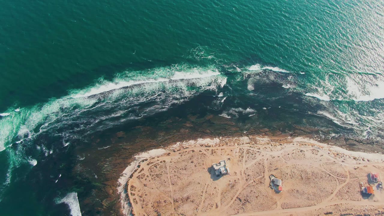 Overhead view of waves breaking over dark nearshore rocks onto a sandy La Bocana beach, with small structures near the lower edge and no visible people.