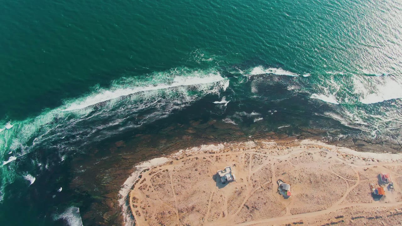Drone view over La Bocana with deep green water, a broken white surf band, dark rocky shallows, sandy beach tracks, and small structures at the lower right, with no visible people.