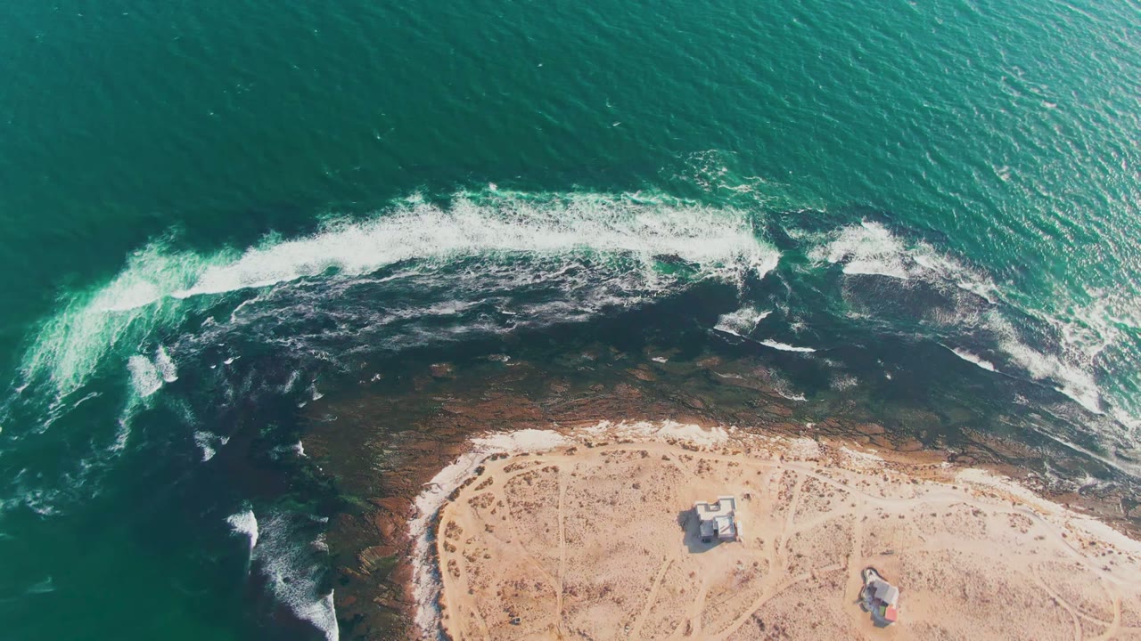 Overhead frame from La Bocana, Baja California Sur, Mexico showing turquoise water, a dark rocky surf line, pale sand, small structures, tracks, and no visible people.