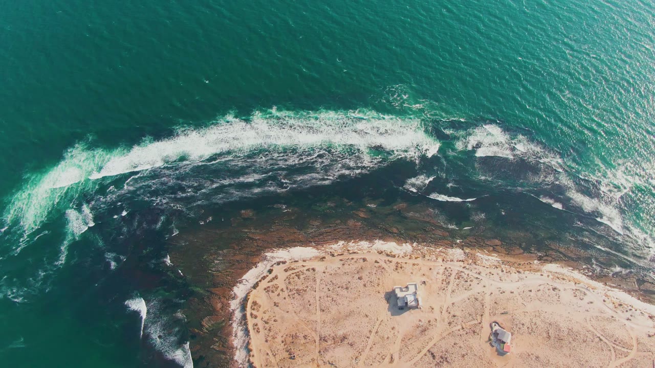 White surf curls across a rocky edge beside pale sand and small beach structures.