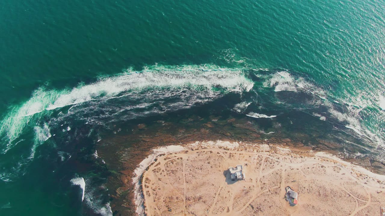 Overhead view in La Bocana showing a bright patch of sunlit sea to the right, surf breaking across dark rocks, sandy ground with tracks, small structures, and no visible people.