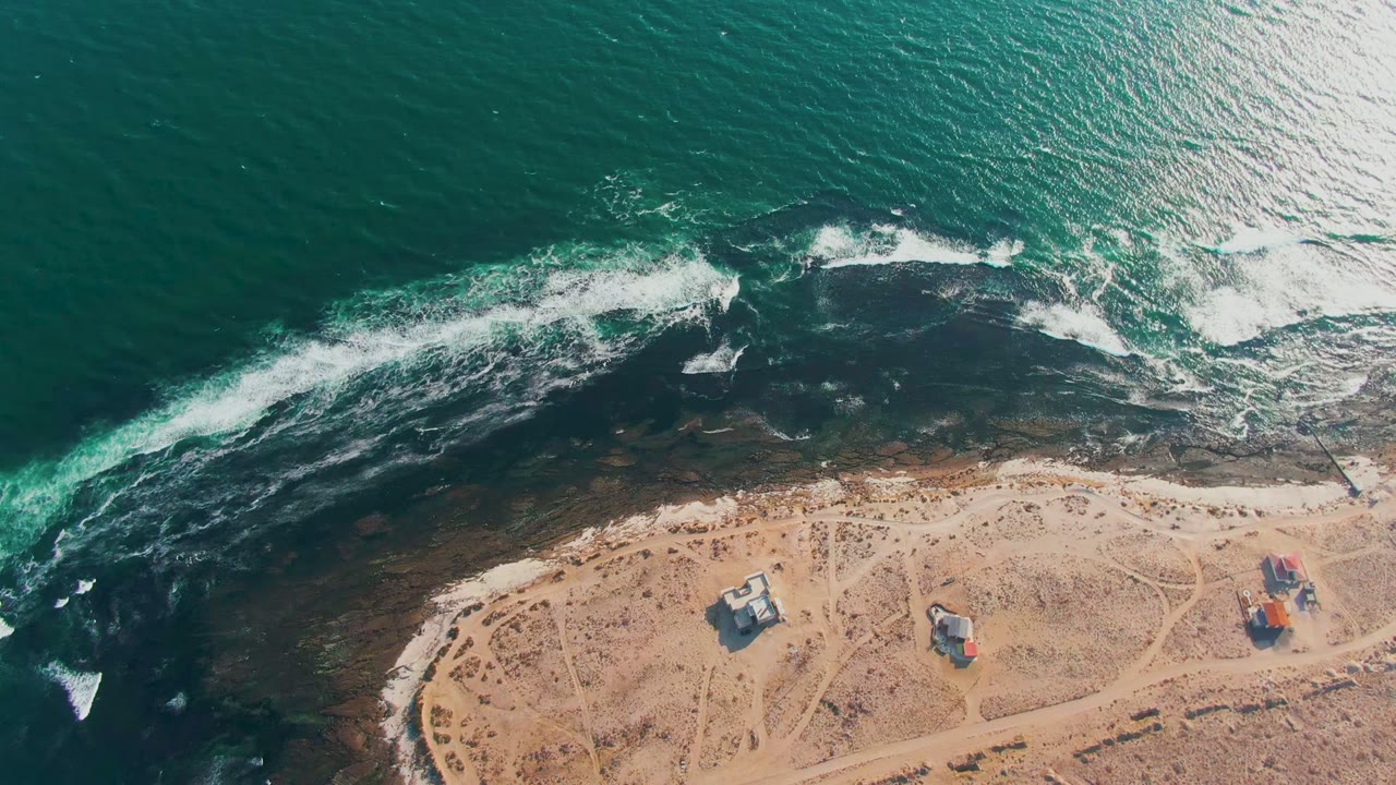 Overhead view of La Bocana surf breaking over dark nearshore rocks beside a tan sandy shoreline with small structures and no visible people.
