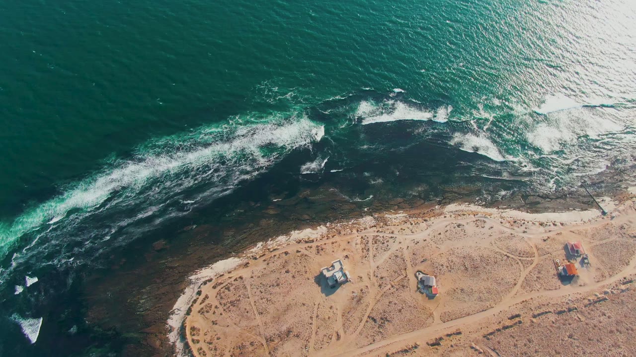 Aerial frame over La Bocana shows green ocean, a broken white surf line, dark shallow rock, and sparse sandy coast with small buildings and no visible people.