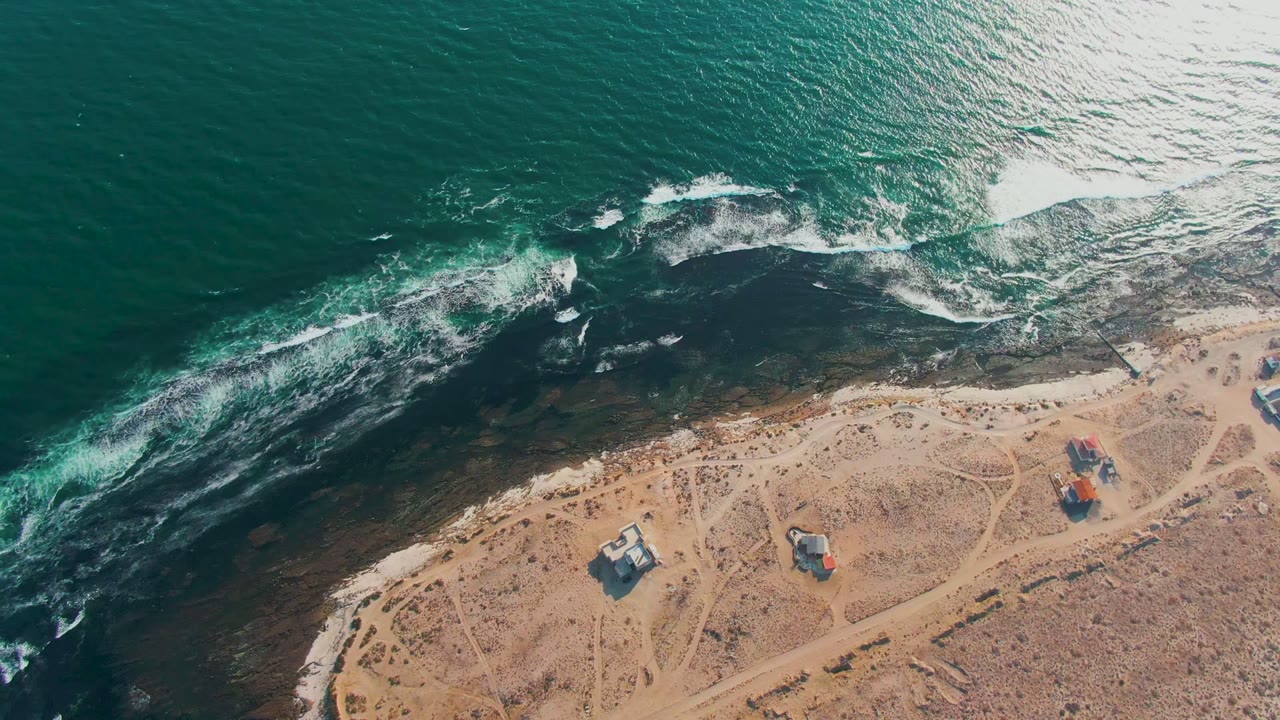 Overhead view of waves breaking along the La Bocana shoreline in Baja California Sur, with dark nearshore rock, sandy terrain, small structures, and no visible people.