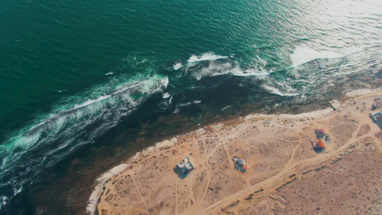 White waves spread across dark rocks near a sandy coast.