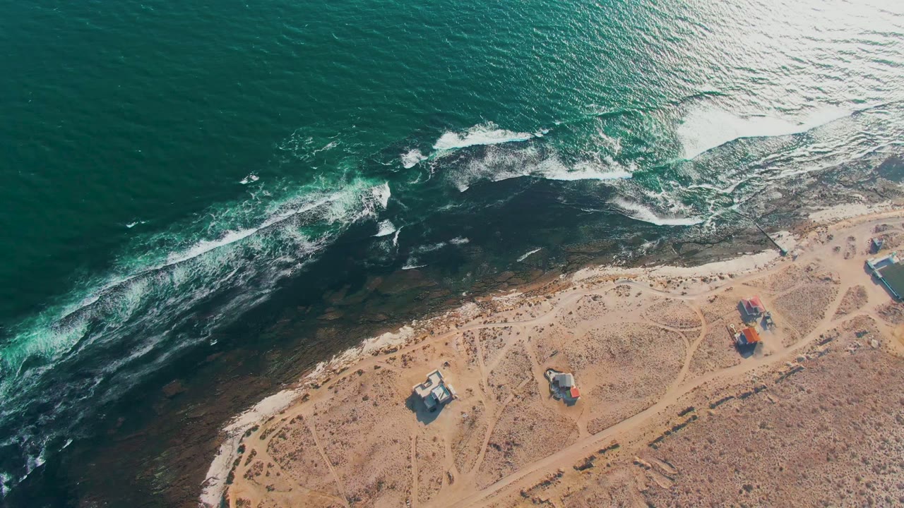 High overhead view of the La Bocana coast as bright surf curves toward the sandy beach, with arid ground, small structures near the shore, and no visible people.