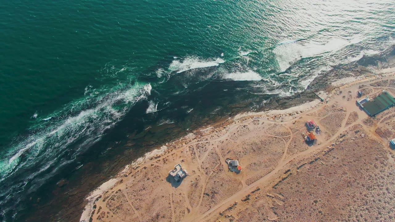 Ocean whitecaps roll toward a dark rocky strip along a sandy shore with huts nearby.