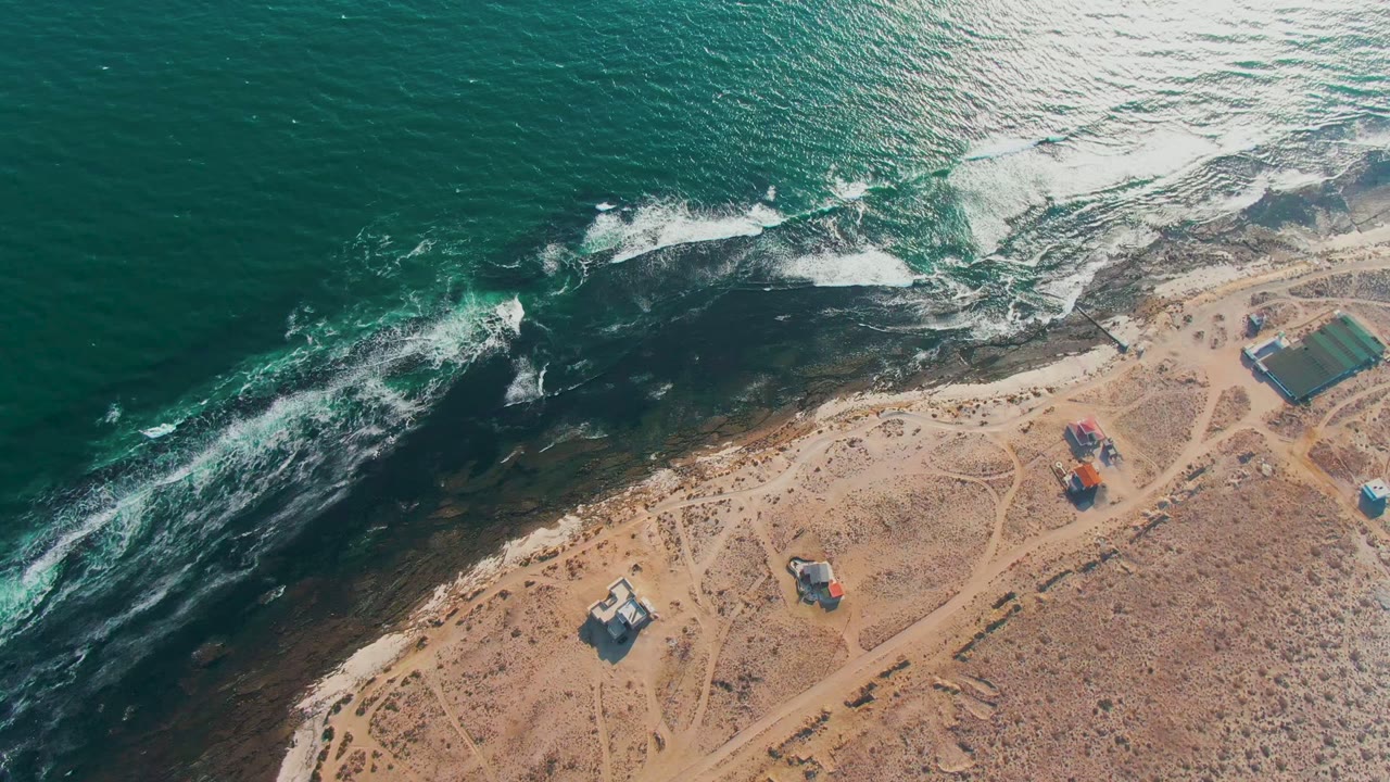 Overhead view of La Bocana with surf breaking along the rocky edge, sandy terrain below, a rectangular coastal structure at right, smaller huts, and no visible people.