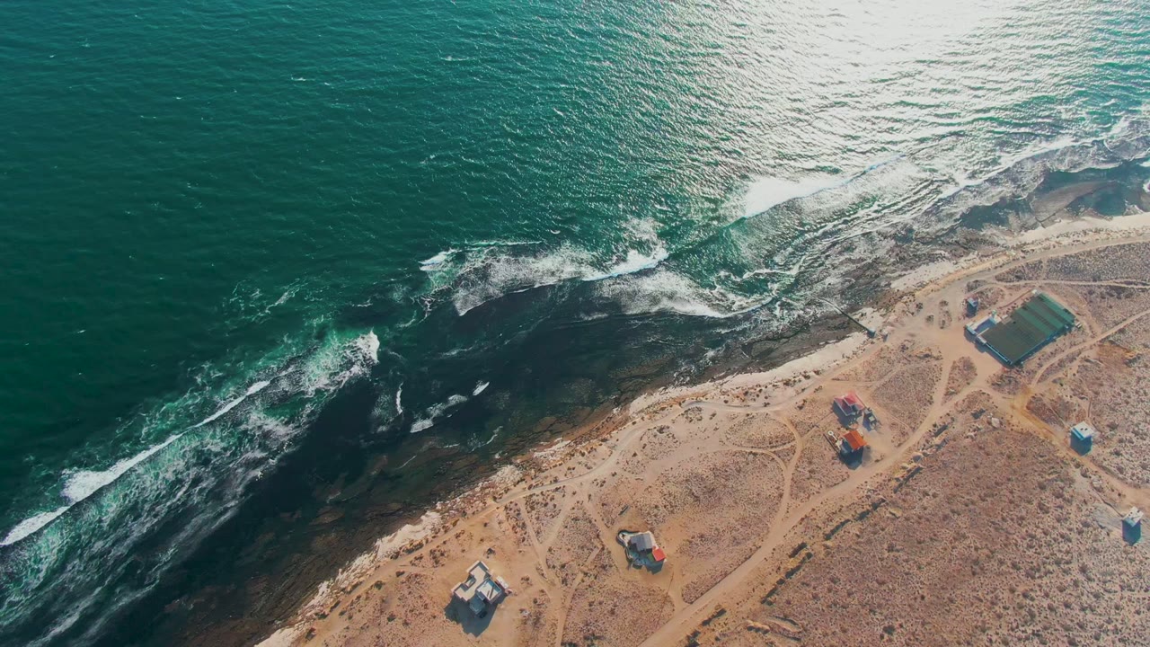 Aerial frame over La Bocana, Baja California Sur showing teal ocean, white surf on dark coastal rocks, sandy tracks, small buildings inland, and no visible people.