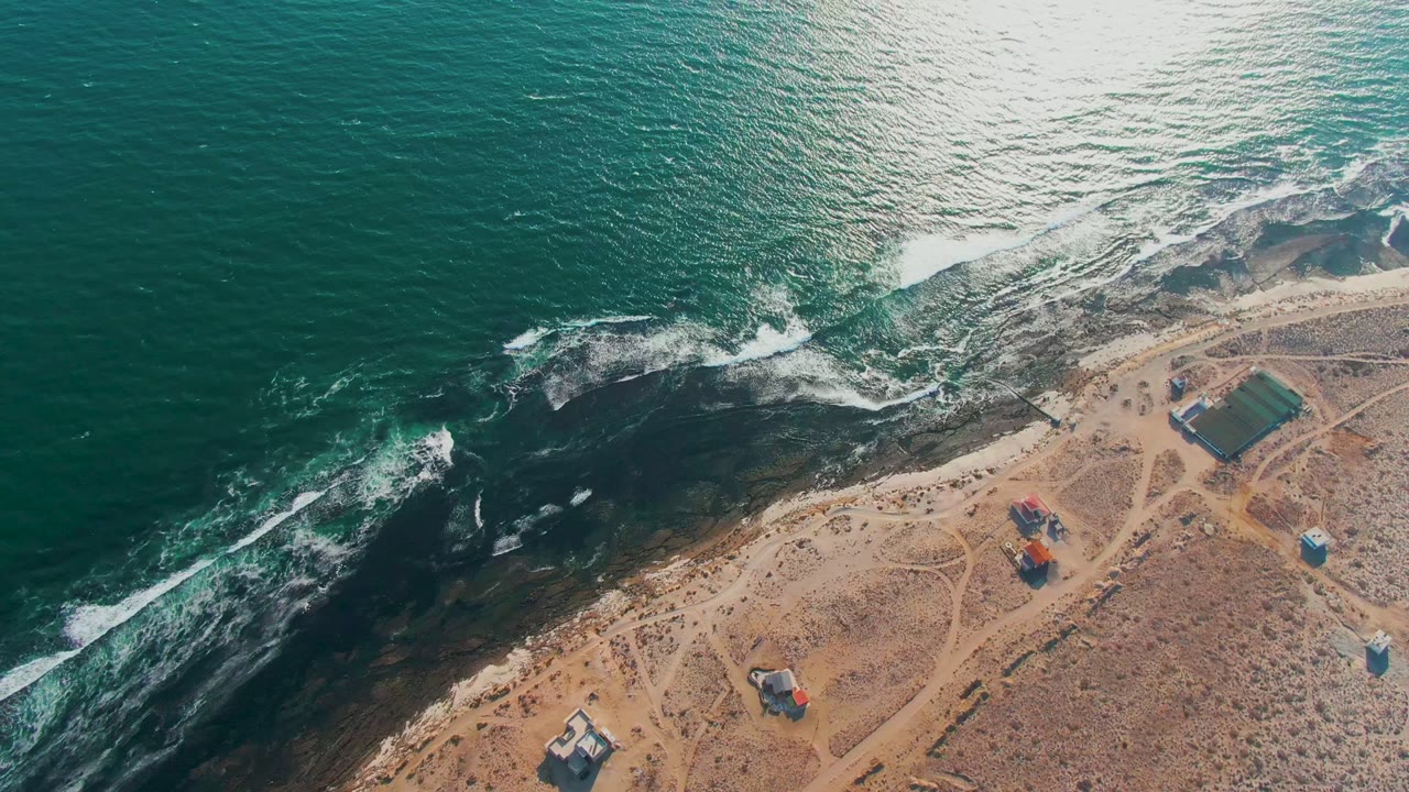 Drone view of surf spreading across dark rocks near sandy coastal land.
