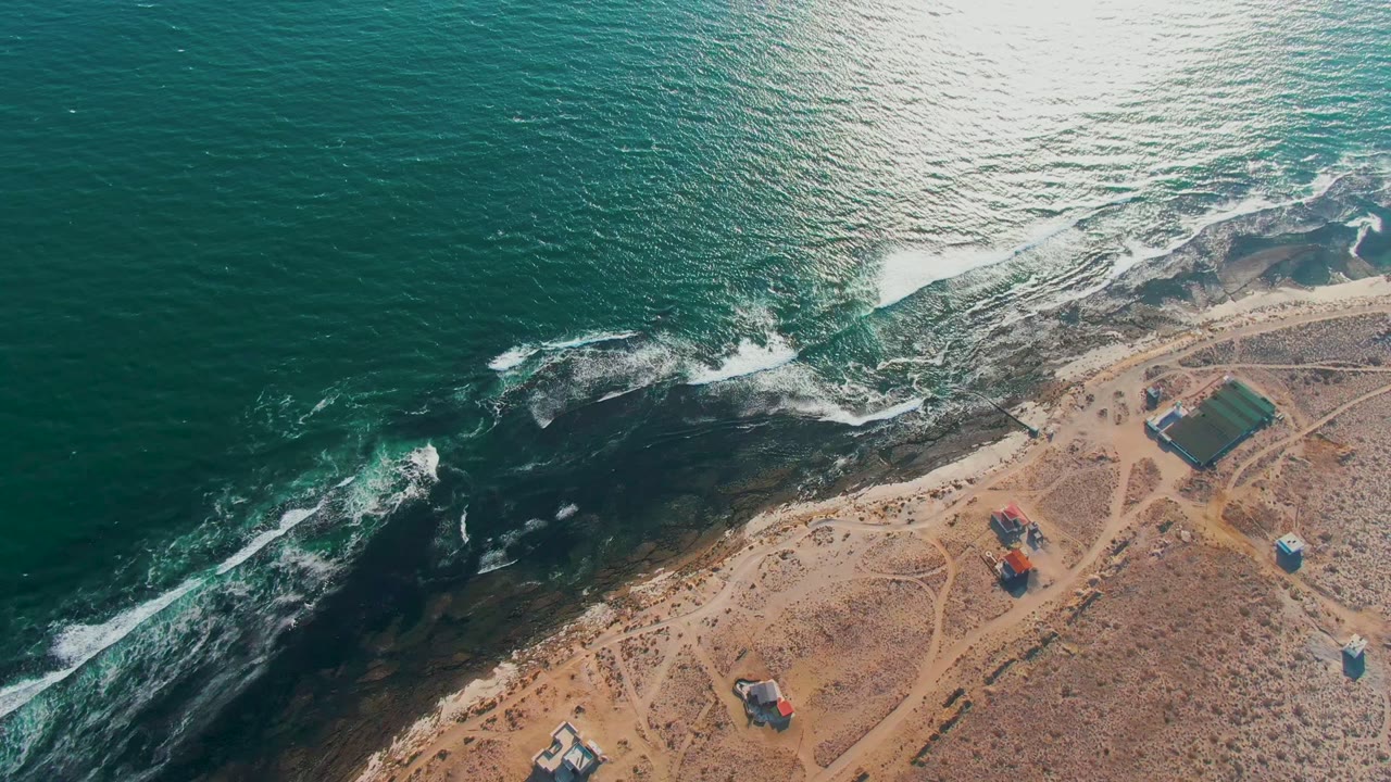Aerial frame of the La Bocana coast with a broad teal water surface, surf breaking diagonally across dark rocks, sparse sandy land, small buildings, and no visible people.