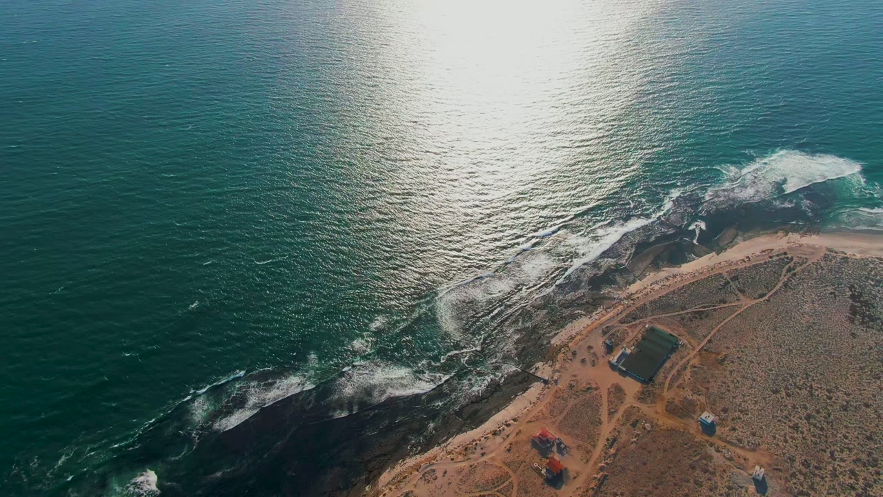 Aerial frame over La Bocana, Baja California Sur, with blue-green ocean, bright sun reflection, white surf on dark rocks, sandy scrub shore, and no visible people.