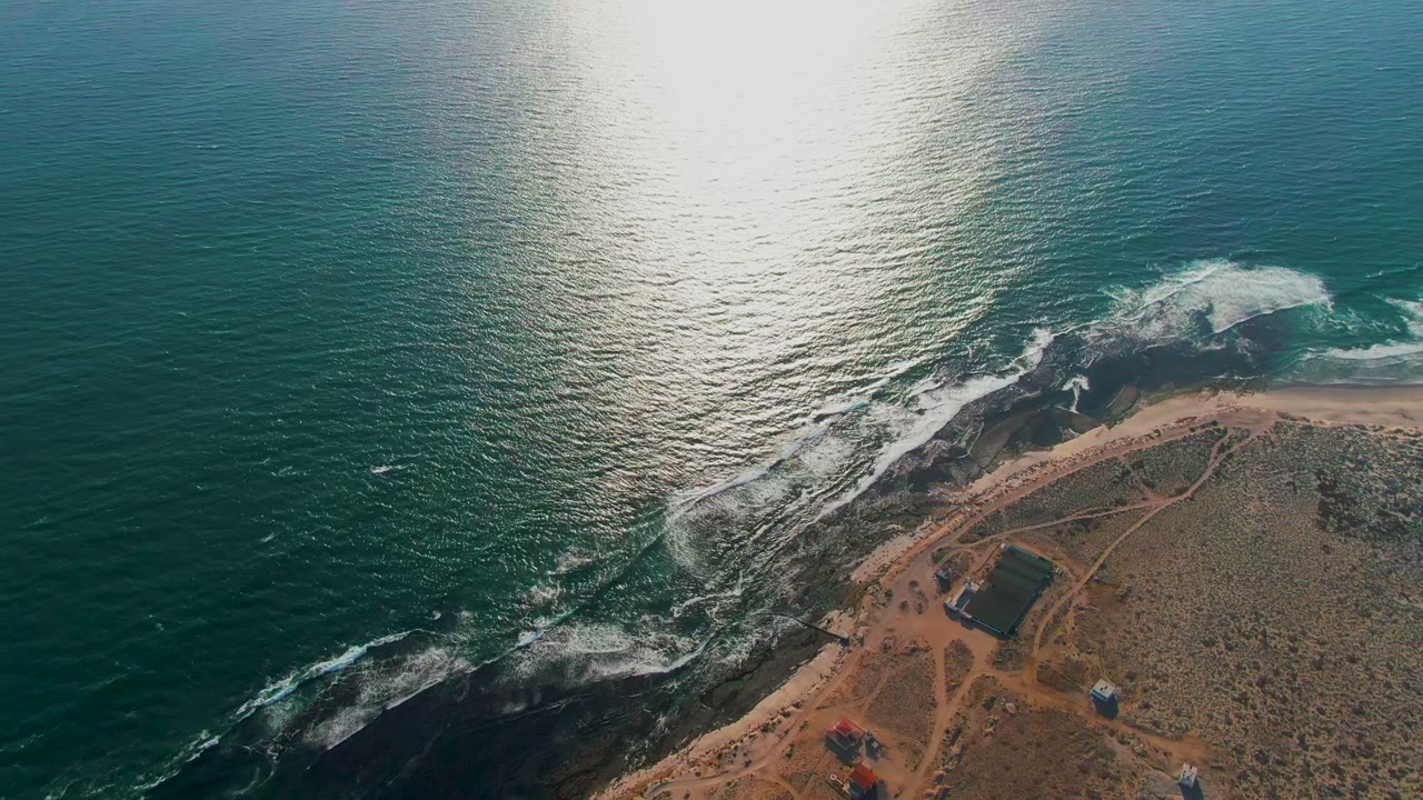 Drone-height frame over La Bocana showing teal ocean, active white breakers, dark rocks, a sandy point with intersecting dirt tracks, and no visible people.