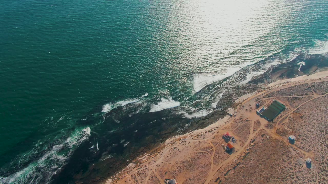 Aerial frame over La Bocana, Baja California Sur, with turquoise water, white foam on dark coastal rocks, sandy tracks, small shore structures, and no visible people.