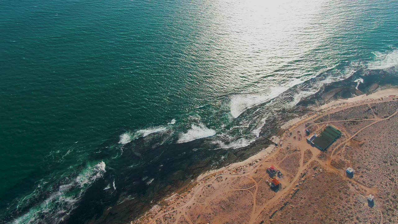 Ocean surf spreads across dark shoreline rocks near sandy coastal tracks.
