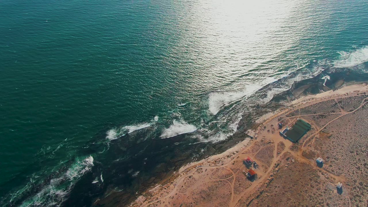 Aerial view in La Bocana with bright sun reflection on the ocean, white waves curling over black rocks, sandy shoreline structures, and no visible people.