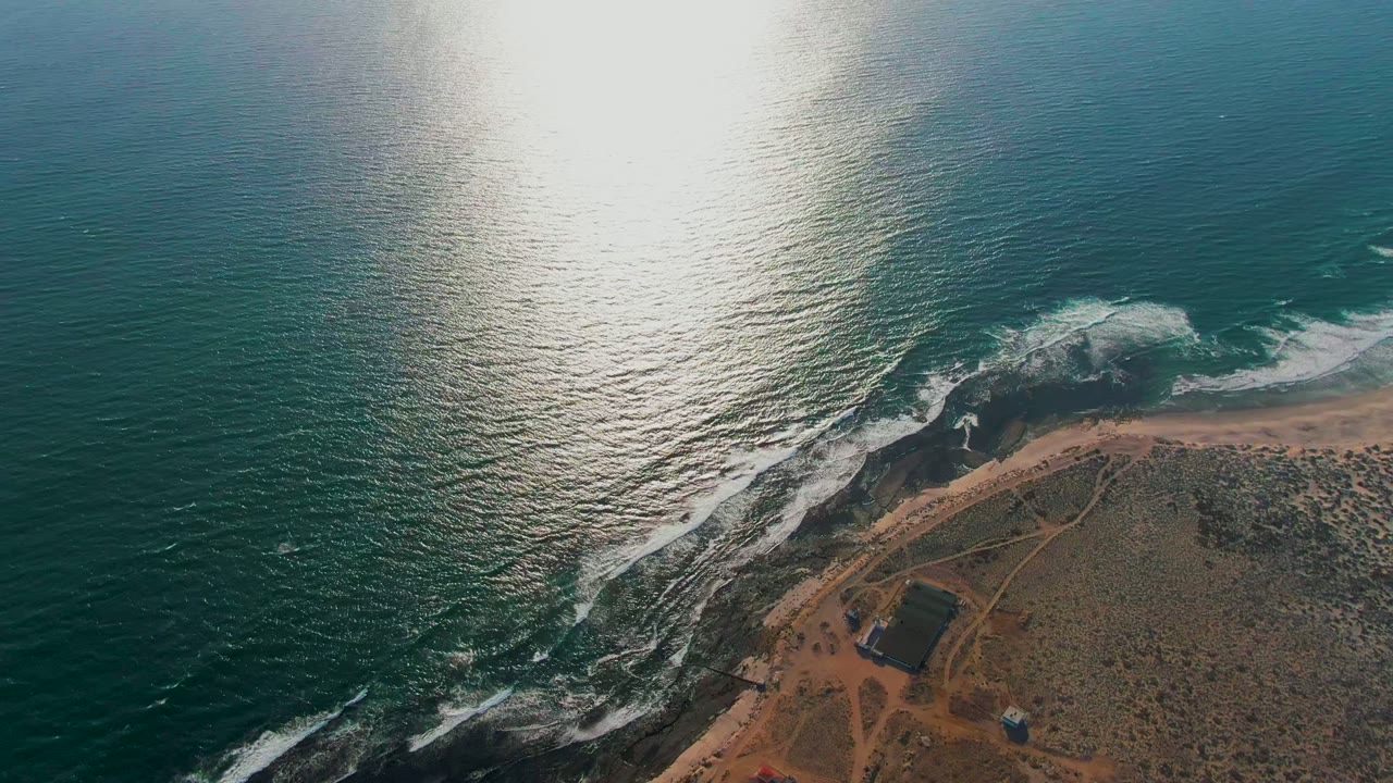Aerial frame over La Bocana, Baja California Sur, with bright sun reflection on teal water, white surf along dark rocks, sandy beach, dry scrubland, a small structure, and no visible people.