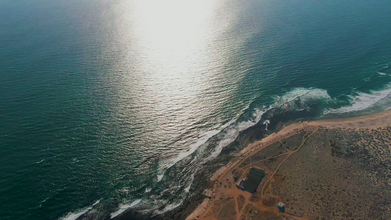 Waves break along dark rocks below a broad patch of reflected sunlight.