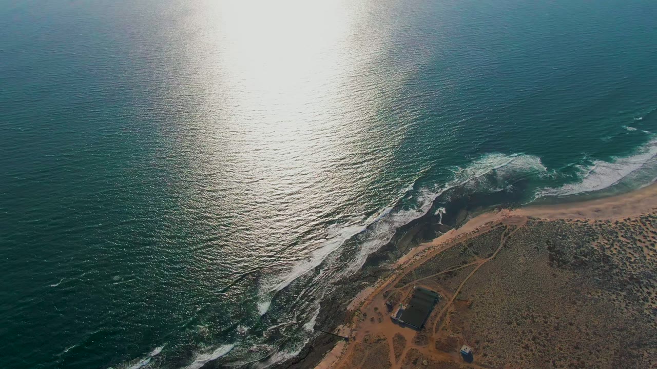 Aerial La Bocana frame with shimmering ocean filling most of the view, a white wave band nearing the beach, dark rock outcrops, scrub-covered land, a dirt path network, and no visible people.
