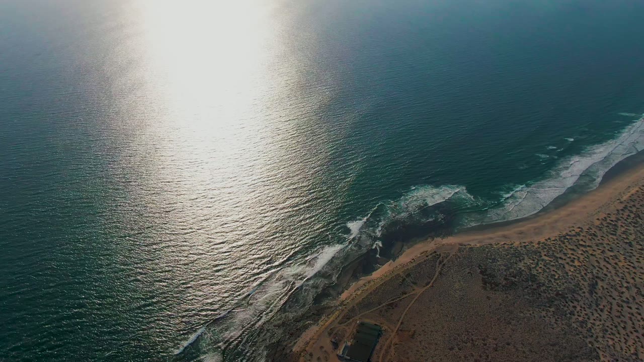 Aerial frame over La Bocana, Baja California Sur, Mexico, showing sunlit teal water, white surf, a curved sandy shore, dark rocks, and no visible people.