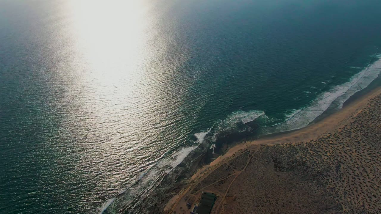 Aerial frame from La Bocana with teal water occupying most of the view, white foam bands along the beach, scrub-covered land at lower right, and no visible people.