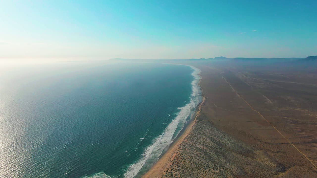 Aerial frame over La Bocana, Baja California Sur with blue ocean on the left, white surf along a sandy beach, arid desert inland, distant mountains, and no visible people.