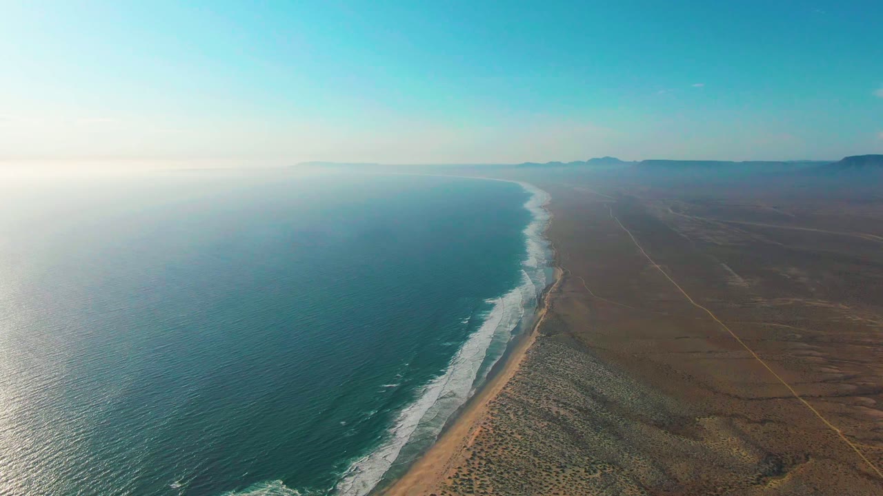Aerial frame over La Bocana, Baja California Sur showing the ocean left of frame, a curling band of surf on the sandy strip, faint inland tracks through dry terrain, distant mountains, and no visible people.