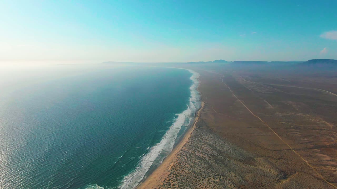 High aerial view over La Bocana, Baja California Sur, with turquoise ocean on the left, white surf along the beach, brown desert terrain to the right, and no visible people.