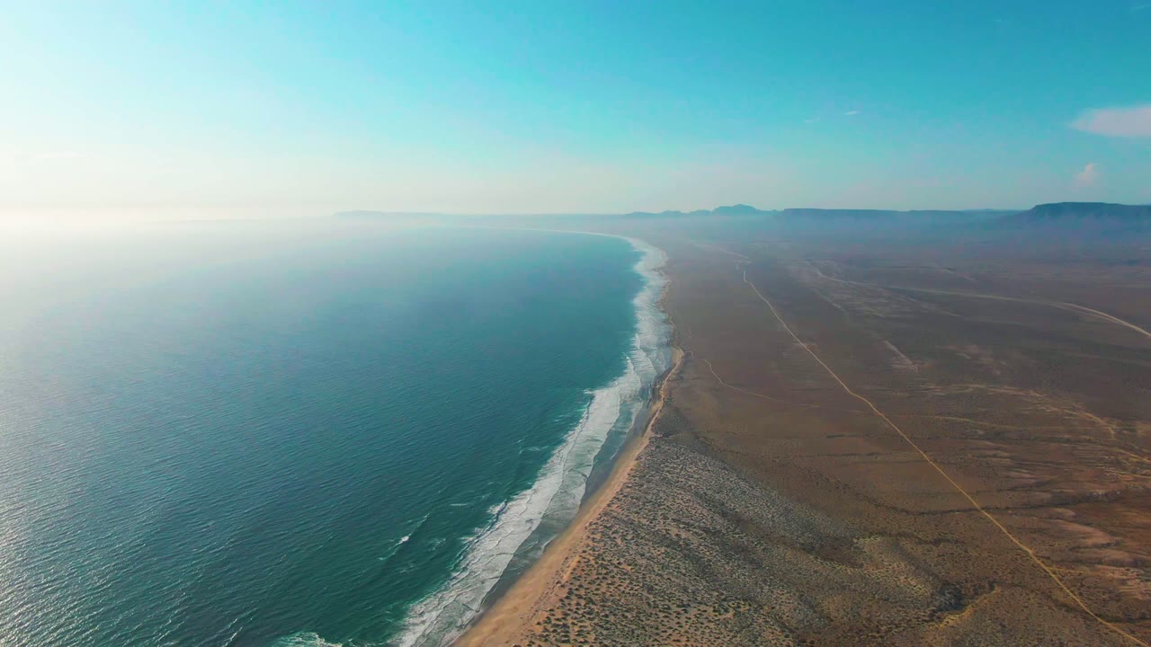 High view of the La Bocana coast with rippled blue-green water, foamy shore break, dry brown land, faint inland tracks, distant mountains, and no visible people.