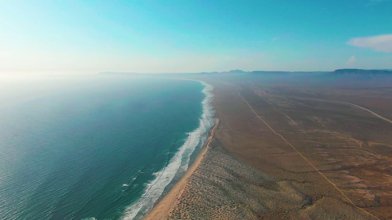 At 0:00, La Bocana's turquoise ocean fills the left side while white surf meets a narrow sandy beach beside dunes and open desert, with no visible people.