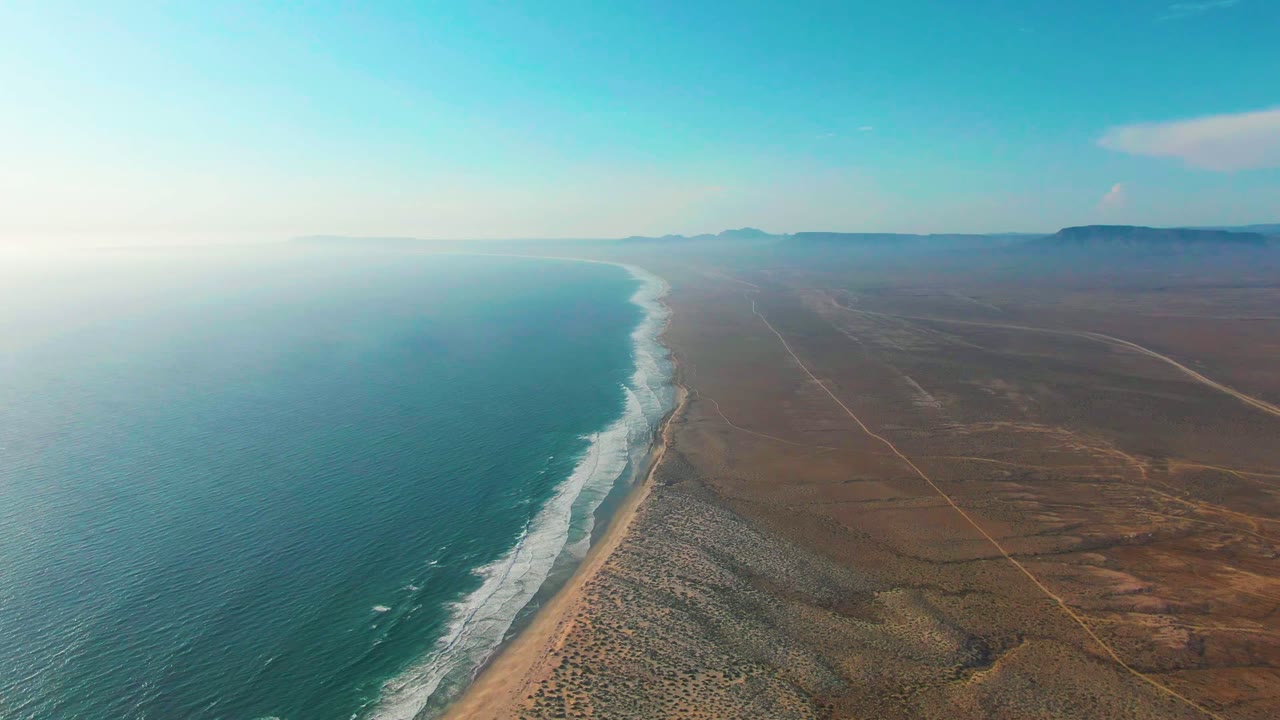 At 0:02, a high aerial view holds the ocean on the left and dry land on the right, with white wave bands along La Bocana's beach and no visible people.
