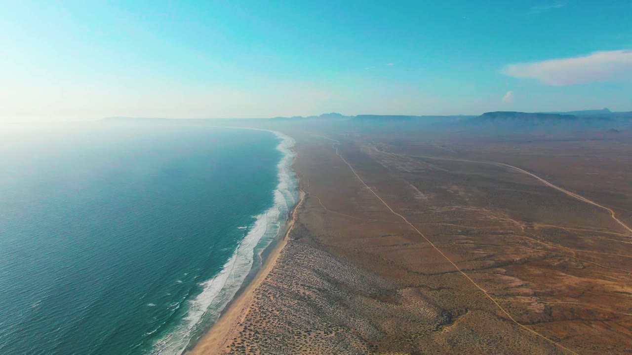 High aerial view over La Bocana, Baja California Sur, with blue ocean on the left, white surf along a sandy rocky shore, brown desert inland, distant hills, and no visible people.
